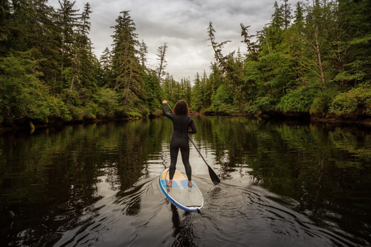 Woman paddlboards on lake amongst evergreens trees.