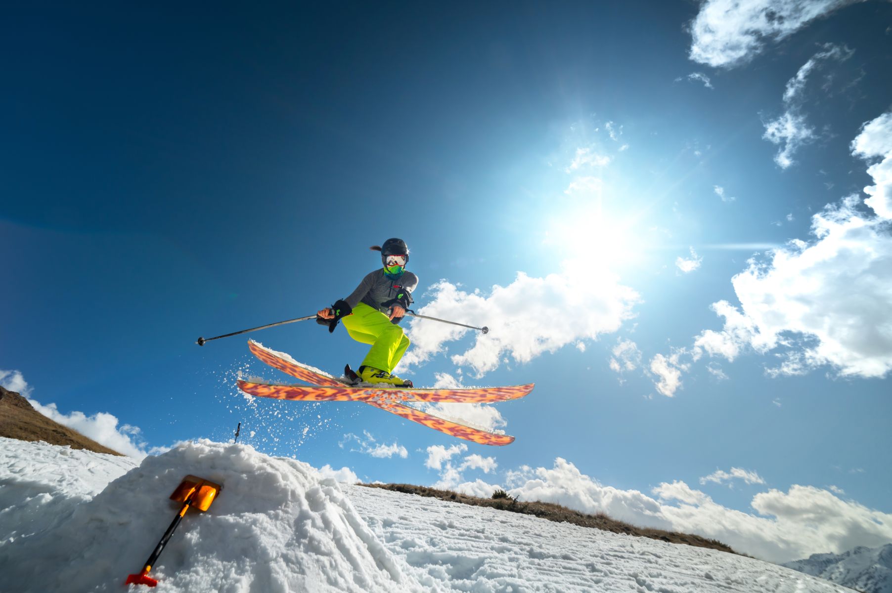 Freestyle skier in neon pants jumping mid-air with bright sun and blue sky behind, skis spread wide apart.