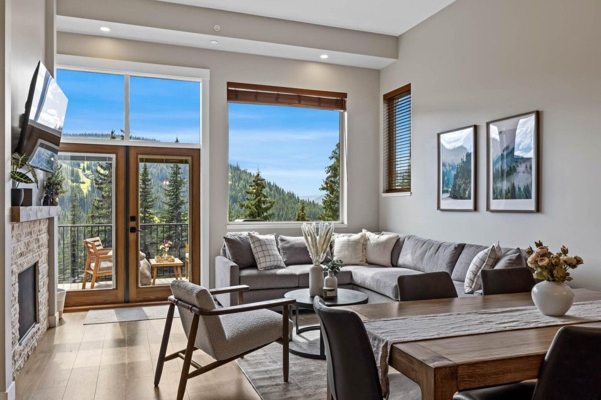 Bright living room with a gray sectional, wooden dining table, and large windows showing mountain scenery outside.