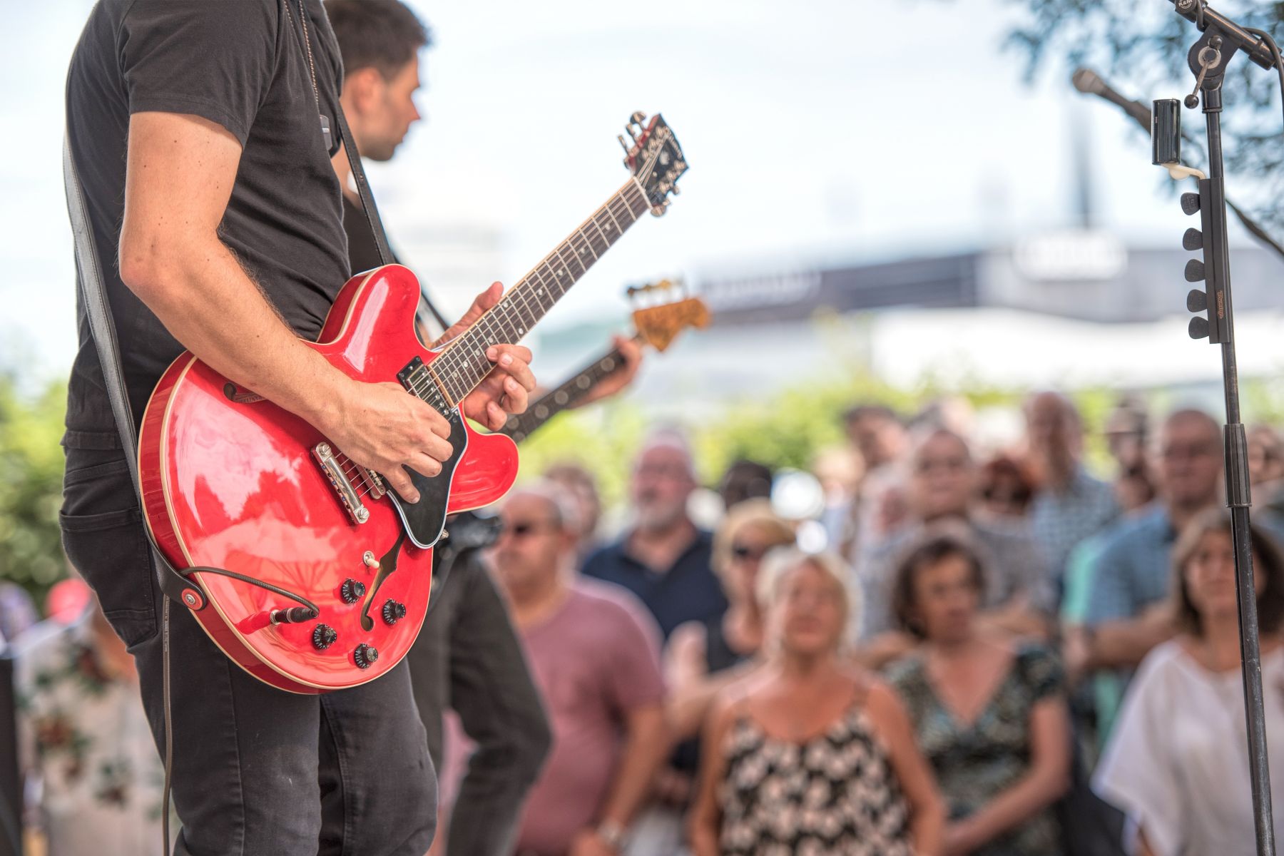 Guitarist in a black shirt playing a red electric guitar on an outdoor stage with a cheering crowd behind him