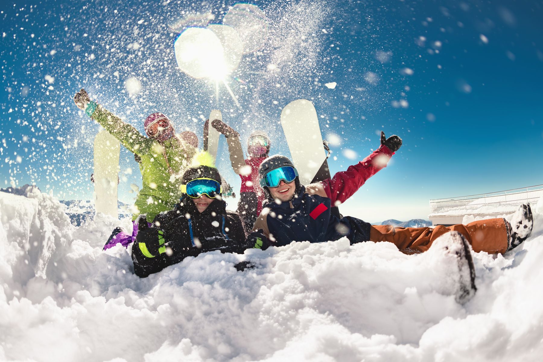 Group of friends in colorful snowsuits cheer with snowboards raised, throwing snow into the air on a sunny slope